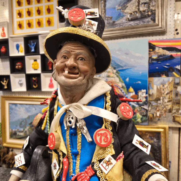 Statuina artigianale napoletana del personaggio della Smorfia con cappello e abiti decorati da numeri e simboli portafortuna, esposta in negozio tra quadri dipinti a mano e ceramiche colorate.