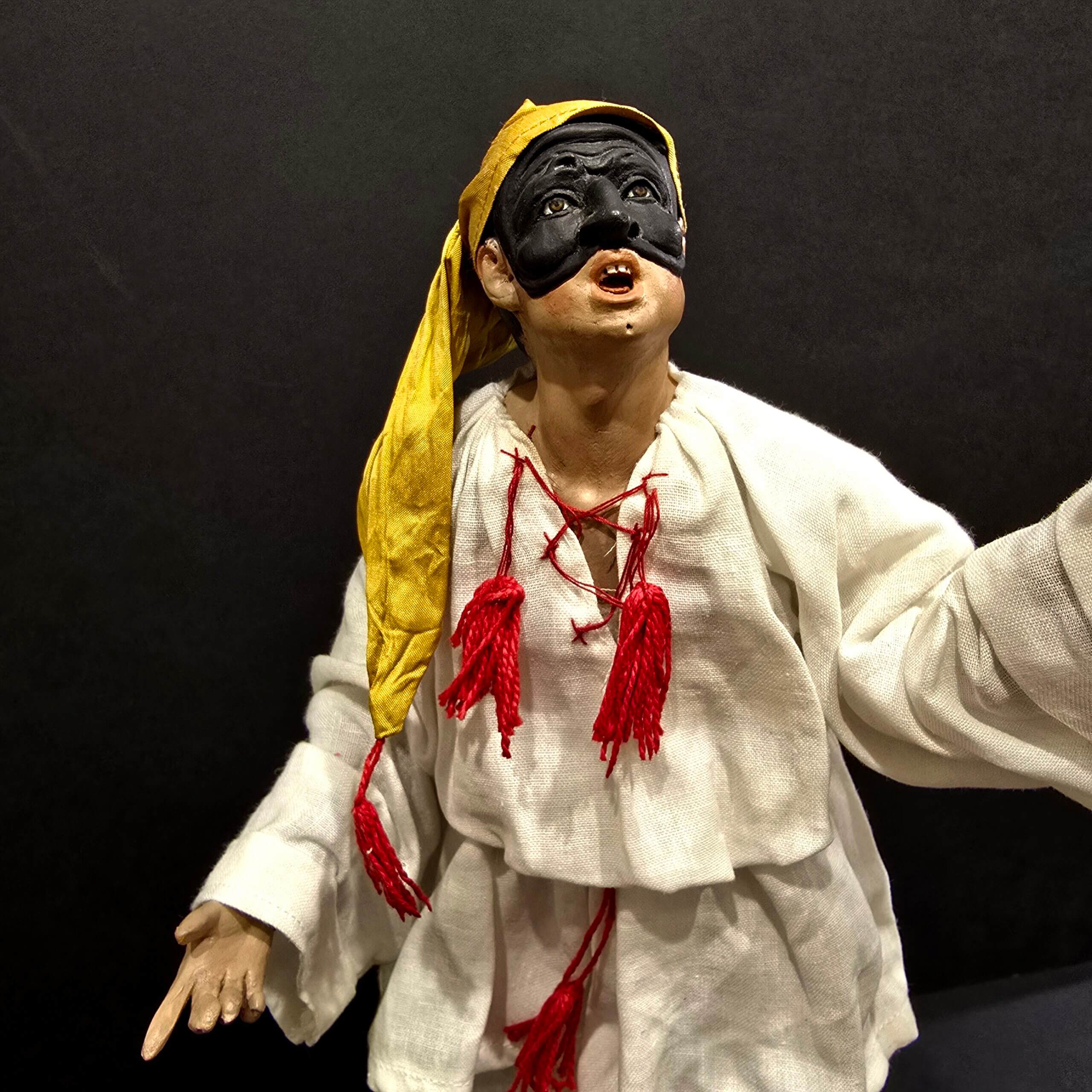 Statua di Pulcinella con berretto giallo e braccia alzate, primo piano con espressione teatrale.