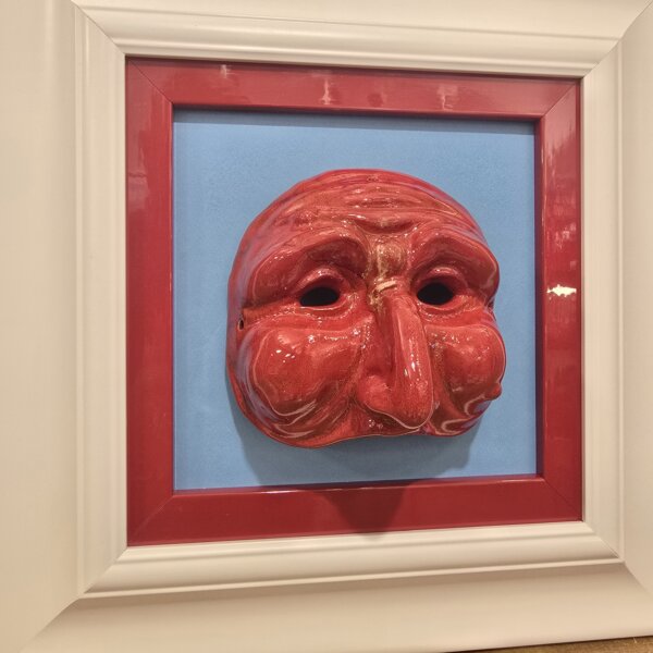 Vista Angolare Maschera Rossa in Cornice Bianca Vista laterale del quadro che evidenzia la tridimensionalità della maschera in ceramica, la finitura lucida dello smalto rosso e la profondità della cornice a cassetta bianca.