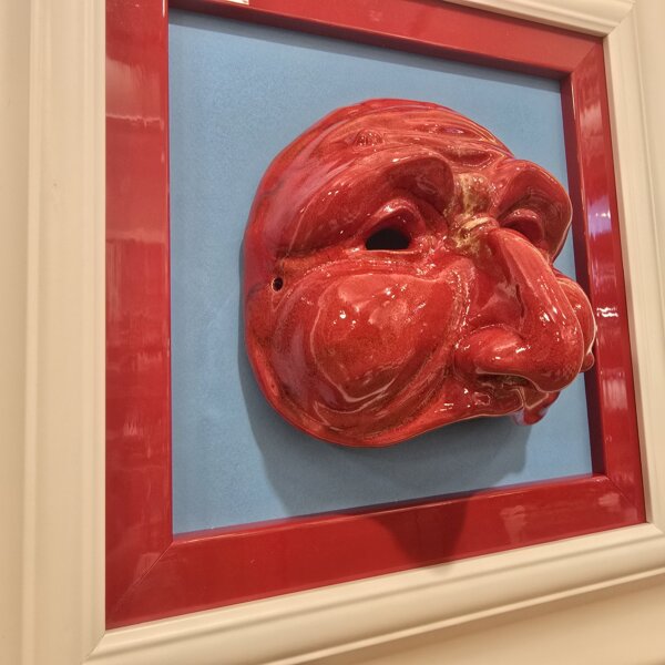 Dettaglio Scultura Ceramica Pulcinella Rosso Primo piano ravvicinato del volto di Pulcinella in ceramica rossa, che mostra i dettagli del naso adunco e delle rughe espressive, in contrasto con lo sfondo celeste.